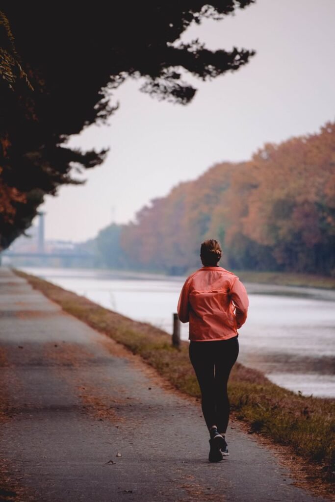 pexels-photo-14104072-14104072 A woman in sportswear jogging along a riverside path surrounded by autumn foliage.